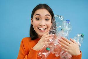 Portrait of cute korean woman, holding two plastic bottles with excited face, recycling her waste, sorting garbage, blue background photo