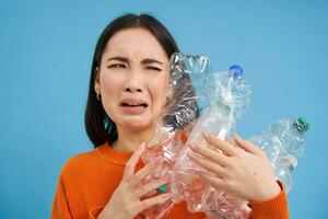 Crying asian girl holding plastic bottles, being upset, recycling, standing over blue background photo