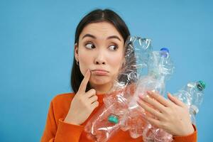 Thoughtful asian woman, holding plastic bottles, looking aside and thinking about recycling, sorting garbage, blue background photo