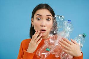 Surprised korean woman, holding plastic bottles, looking shocked at camera, concept of environment and recycling, blue background photo