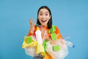 Surprised asian woman, sorting garbage, recycling, looks excited, carries plastic waste, blue background photo