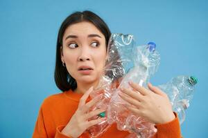 Portrait of asian girl with scared face, holding piles of plastic bottles for recycling and stares to the right with shocked emotion, blue backgrond photo