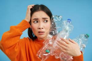 Portrait of asian girl with scared face, holding piles of plastic bottles for recycling and stares to the right with shocked emotion, blue backgrond photo