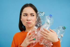 Annoyed korean woman looking with blaming face, holding plastic bottles, standing over blue studio background photo