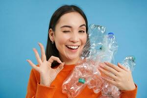 Recycling is great. Smiling young woman, holding bottles, shows okay sign, approves eco green lifestyle, sorting garbage, blue background photo