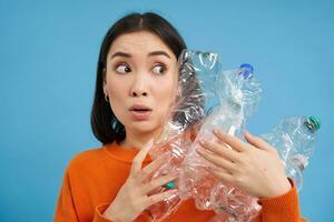 Portrait of asian girl with scared face, holding piles of plastic bottles for recycling and stares to the right with shocked emotion, blue backgrond photo