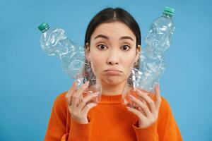 Portrait of sad asian woman with plastic bottles, upset by lack of recycling centers, blue background photo