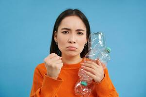 Angry asian woman holding two plastic bottles with frowning face, girl with eco green lifestyle, urges to recycle waste, blue background photo