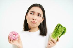 Healthy clean detox eating concept. Vegetarian, vegan, raw concept. Sad asian girl on diet holds doughnut and vegetable, wants to taste donnut, white background photo