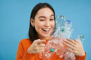 usted lata reciclar. entusiasta joven asiático mujer, guiños y puntos a cámara, muestra el plastico botellas, alienta para reciclaje y clasificación desperdiciar, azul antecedentes foto