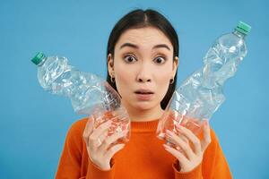 Portrait of girl with two plastic bottles, looks confused, tried to recycle, sorting garbage, isolated on blue background photo