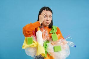 Shocked asian woman, looks embarrassed at something horrible, holding plastic garbage, sorting waste for recycling, blue background photo