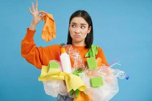 Young woman holds plastic garbage, looks at glove with shocked face, stinky waste, sorting litter for recycling, blue background photo