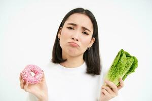 Healthy clean detox eating concept. Vegetarian, vegan, raw concept. Sad asian girl on diet holds doughnut and vegetable, wants to taste donnut, white background photo