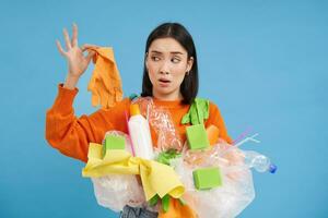 Young woman holds plastic garbage, looks at glove with shocked face, stinky waste, sorting litter for recycling, blue background photo