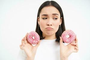 cerca arriba retrato de triste asiático mujer, trastornado siendo en dieta, demostración dos vidriado rosado donas, tentado a comer basura alimento, blanco antecedentes foto