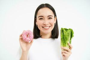 Healthy lifestyle concept. Happy smiling asian woman, showing one glazed doughnut and cabbage, giving choice, white background photo