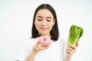 Healthy clean detox eating concept. Vegetarian, vegan, raw concept. Smiling korean girl eats delicious doughnut, holds cabbage, enjoys junk food, white background photo