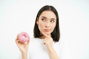 imagen de asiático mujer sostiene rosquilla y piensa, hace decisión, soportes terminado blanco estudio antecedentes foto