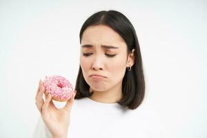 llorando asiático mujer demostración sabroso donuts y sollozando, siendo en sano dieta, hipocresía comer basura alimento, blanco antecedentes foto