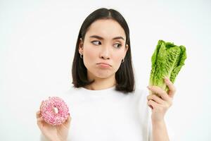 sano estilo de vida concepto. joven mujer decidiendo Entre sano verduras, repollo y delicioso vidriado donas, blanco antecedentes foto