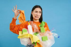 Korean woman looks at stinky latex glove, holds plastic garbage, cleans house and recycle, blue background photo