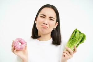 Healthy clean detox eating concept. Vegetarian, vegan, raw concept. Sad asian girl on diet holds doughnut and vegetable, wants to taste donnut, white background photo