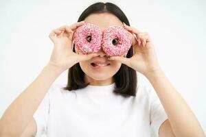 imagen de gracioso joven mujer, hace lentes con dos rosado vidriado donas, mirando mediante rosquilla agujeros y sonriente, aislado en blanco antecedentes foto