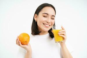 Woman drinking fresh juice, holding orange fruit and smiling, feeling healthy and hydrated, white studio background photo