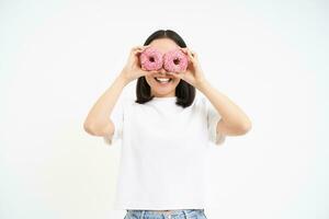 retrato de contento coreano mujer, hace lentes desde dos sabroso vidriado donas, sonriente y teniendo divertido, blanco antecedentes foto