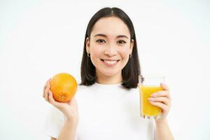 cerca arriba retrato de morena chica, 25 años, participación Fresco vaso de jugo y naranja fruta, sonriente, come sano alimento, blanco antecedentes foto