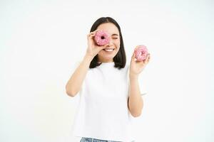 Portrait of happy korean woman, makes glasses from two tasty glazed doughnuts, smiling and having fun, white background photo