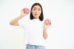 imagen de asiático niña sostiene dos donas, pensando de rotura su dieta, mira con duda a donas, blanco antecedentes foto