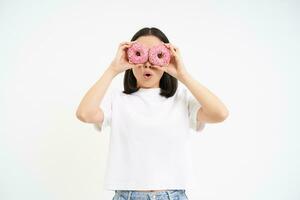 imagen de gracioso joven mujer, hace lentes con dos rosado vidriado donas, mirando mediante rosquilla agujeros y sonriente, aislado en blanco antecedentes foto