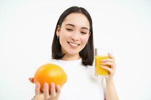 Portrait of smiling brunette woman, squeeze one glass of fresh juice from orange and looking happy, has healthy drink, white background photo