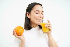 Close up portrait of asian woman, drinking glass of orange juice with delight and happy smile, white studio background photo