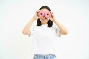 Image of funny young woman, makes glasses with two pink glazed doughnuts, looking through donnut holes and smiling, isolated on white background photo