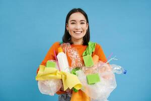 Happy girl looks at plastic garbage, sorting her waste for recycling station, smiling excited, blue background. Environment and ecology concept photo