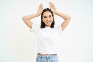 Funny asian woman, shows bunny ears gesture, posing cute against white background photo