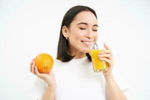 Close up portrait of asian woman, drinking glass of orange juice with delight and happy smile, white studio background photo