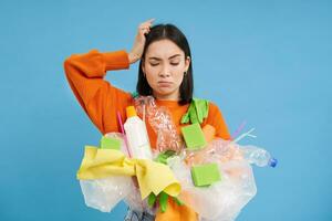 Confused Korean woman tries to go green, recycle plastic household items, holding bottles and gloves, does recycling, blue background photo