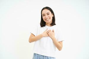 retrato de sonriente, despreocupado asiático chica, sostiene manos en corazón y risas, soportes aislado en blanco antecedentes foto