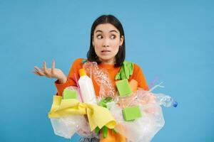 Korean girl with plastic garbage, shrugs shoulders, doesnt know how to recycle, sorting her waste for recycling station, blue background photo