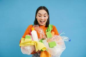 Happy girl looks at plastic garbage, sorting her waste for recycling station, smiling excited, blue background. Environment and ecology concept photo