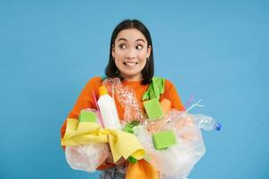 Enthusiastic asian woman holding non sorted plastic garbage, recycling materials with happy face, standing over blue background photo
