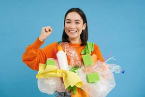 Joyful asian girl holds recycable plastic waste and cheering, go green, cleaning household from garbage, sorting and recycle, blue background photo