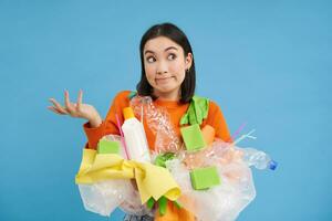 Korean girl with plastic garbage, shrugs shoulders, doesnt know how to recycle, sorting her waste for recycling station, blue background photo