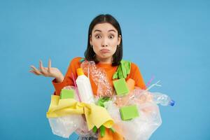 Korean girl with plastic garbage, shrugs shoulders, doesnt know how to recycle, sorting her waste for recycling station, blue background photo