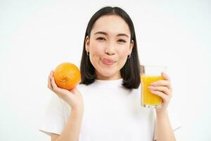 Close up of smiling brunette girl, drinks fresh juice, holds orange fruit, white studio background photo