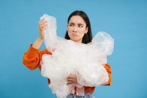 Young asian woman with plastic waste, looking bothered by recycling, standing over blue background photo